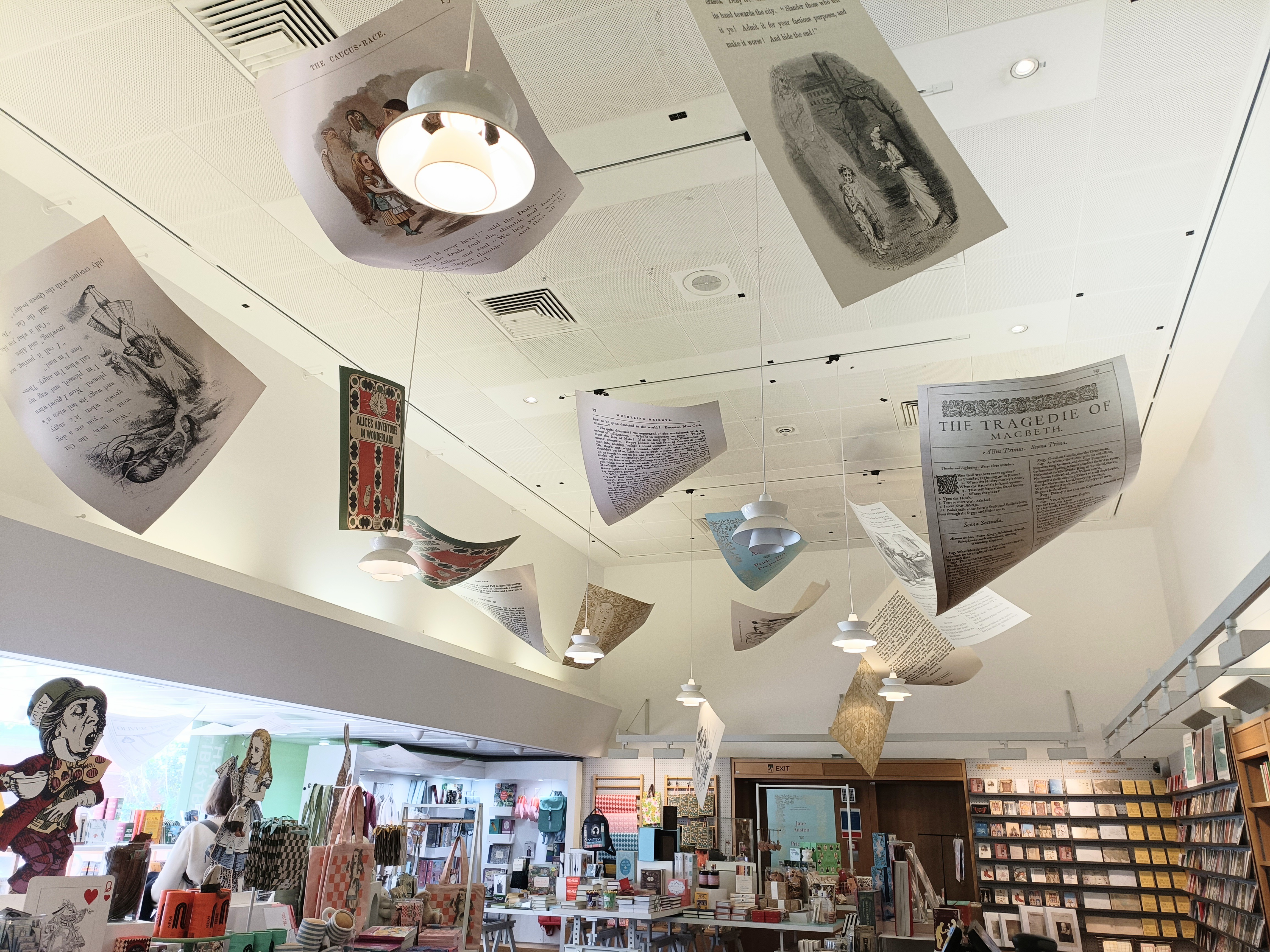 A picture of an indoor ceiling, from which hangs art pages made to look as though they are pages of books. Below there are walls lined with bookshelves and further art.
