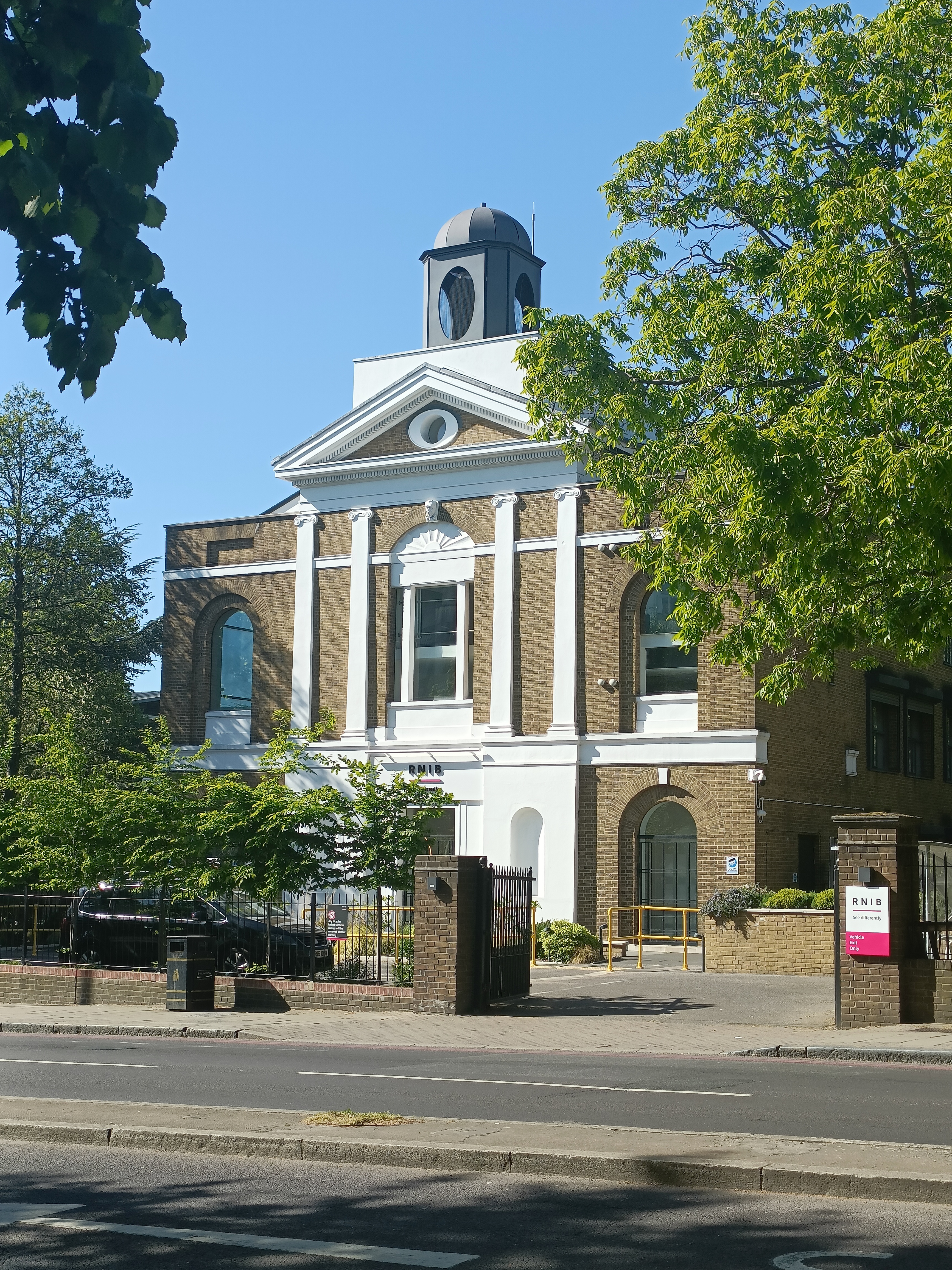 An image of the outside of the Heritage Centre- a brown brick building with a white entrance & pillars. In front of the building are a number of green trees, and behind is a blue sky.